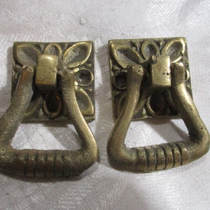 May include: Two antique brass door or cabinet pulls with ornate floral designs and a triangular loop handle.