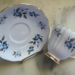 May include: A white teacup and saucer with a blue and gray floral pattern. The saucer has a gold rim. The teacup has a gold rim and a fluted design.