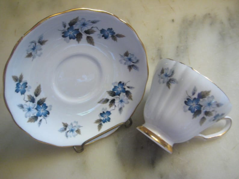 May include: A white teacup and saucer with a blue and gray floral pattern. The saucer has a gold rim. The teacup has a gold rim and a fluted design.