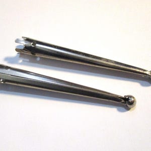 May include: Two silver-colored metal tools with a pointed end and a small ball on the opposite end. The tools are angled and have a three-pronged fork-like design at the pointed end. The tools are on a white background.