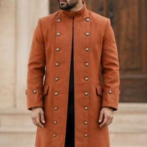 May include: A long, burnt orange coat featuring a high collar and a double-breasted front closure with two vertical rows of metallic buttons. The coat has long sleeves, structured shoulders, and two side pockets, worn over black trousers and black boots.
