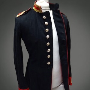 May include: A navy blue military-style jacket with a high collar and gold buttons. The jacket features red trim along the edges, cuffs, and shoulder epaulets. Gold stripes adorn the cuffs. The jacket is displayed on a mannequin.