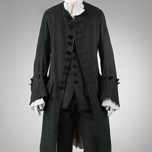 Puede incluir: Un traje de época de tres piezas de color gris oscuro. El abrigo largo tiene botones negros y puños decorativos. También se incluye un chaleco a juego y calzones hasta la rodilla. Una camisa blanca con volantes es visible en el cuello y los puños. El conjunto se exhibe en un maniquí.