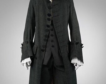 May include: A dark gray, three-piece period costume. The long coat has black buttons and decorative cuffs. A matching vest and knee-length breeches are also included. A white ruffled shirt is visible at the neck and cuffs. The outfit is displayed on a mannequin.
