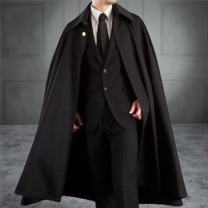 Puede incluir: Una capa negra con un traje a juego. La capa tiene un cuello ancho y un botón dorado. El traje incluye una camisa blanca con cuello y una corbata negra. El conjunto es formal y elegante.