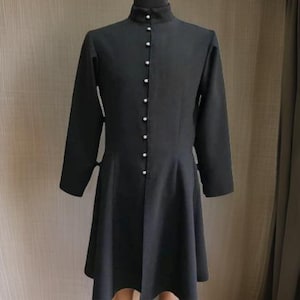May include: A long-sleeved black dress with a high collar and a row of white buttons down the front. The dress has a fitted bodice and a flared skirt, and is made of a smooth, dark fabric.