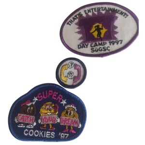 May include: Three embroidered patches. One oval patch with the text "THAT'S ENTERTAINMENT!" and "DAY CAMP 1997 SGGSC". A round patch with a phone graphic. A blue patch with the text "SUPER COOKIES '97" and cartoon cookie characters holding signs that say "CATCH YOUR DREAM".