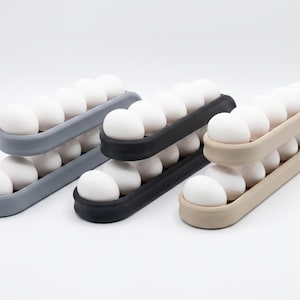 May include: Three tiered egg storage racks in gray, black, and beige. Each rack features two levels of curved channels designed to hold multiple white eggs securely in place. The structures are made of a smooth, matte material with rounded edges.