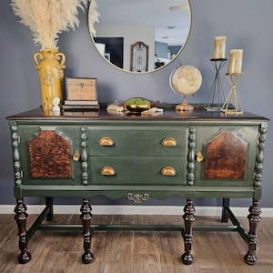 May include: A green and brown antique buffet with gold hardware. The buffet has two drawers and two cabinet doors with decorative wood panels. A round gold mirror hangs above the buffet, which is decorated with a globe, candles, and a yellow vase.