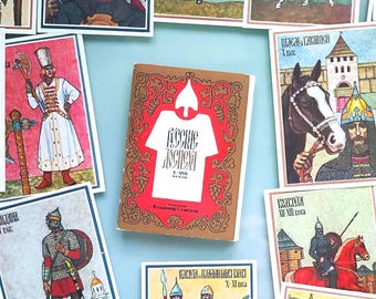 May include: A collection of vintage cards and a book featuring illustrations of medieval warriors, armor, and weaponry. The cards and book have a red and brown color scheme, with text in a foreign language. The cards are scattered on a light blue surface.