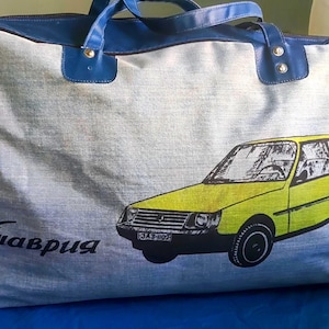 Könnte beinhalten: Eine rechteckige Reisetasche mit einem hellgrauen, strukturierten Stoffkörper und dunkelblauen Besätzen. Die Tasche verfügt über zwei dunkelblaue Griffe und einen Grafikdruck eines gelben Schrägheckautos mit dem kyrillischen Text "Таврия" in Schwarz.