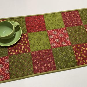 Puede incluir: Una taza y un platillo verdes descansan sobre un camino de mesa de patchwork. El camino de mesa presenta un patr&oacute;n repetitivo de cuadrados rojos y verdes con dise&ntilde;os florales y de cachemira. Los bordes est&aacute;n ribeteados con un borde verde.
