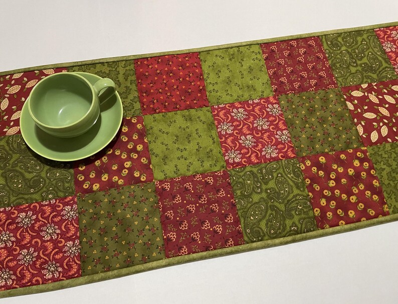 Puede incluir: Una taza y un platillo verdes descansan sobre un camino de mesa de patchwork. El camino de mesa presenta un patr&oacute;n repetitivo de cuadrados rojos y verdes con dise&ntilde;os florales y de cachemira. Los bordes est&aacute;n ribeteados con un borde verde.