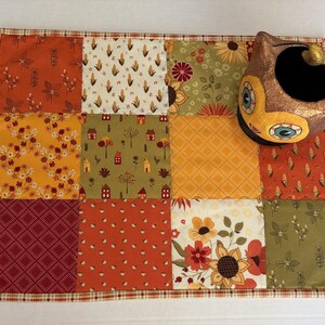 Peut inclure: Un set de table rectangulaire en tissu compos&eacute; de douze carr&eacute;s en patchwork aux motifs automnaux dans des tons orange, jaune, vert et bordeaux. Un r&eacute;cipient en c&eacute;ramique en forme de hibou &agrave; la finition bronze m&eacute;tallique est pos&eacute; en haut &agrave; droite.