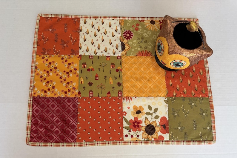 Peut inclure: Un set de table rectangulaire en tissu compos&eacute; de douze carr&eacute;s en patchwork aux motifs automnaux dans des tons orange, jaune, vert et bordeaux. Un r&eacute;cipient en c&eacute;ramique en forme de hibou &agrave; la finition bronze m&eacute;tallique est pos&eacute; en haut &agrave; droite.