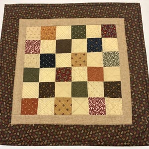 May include: A square patchwork quilt featuring a grid of small fabric squares in various earth tones including cream, brown, olive green, and deep red. The center is framed by a textured tan border and an outer border with a small leaf pattern.