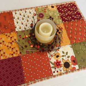 Peut inclure: Un set de table matelass&eacute; rectangulaire pr&eacute;sentant un motif en patchwork de tissus aux couleurs automnales dans des tons orange, jaune, rouge et vert olive. Un bougeoir en verre avec une bougie pilier blanche repose sur une couronne de brindilles.