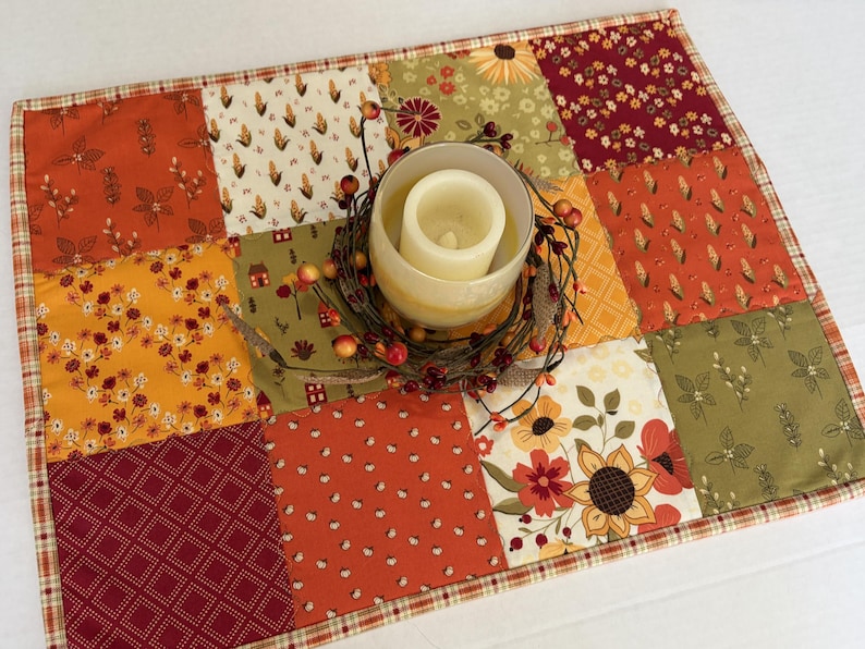 Peut inclure: Un set de table matelass&eacute; rectangulaire pr&eacute;sentant un motif en patchwork de tissus aux couleurs automnales dans des tons orange, jaune, rouge et vert olive. Un bougeoir en verre avec une bougie pilier blanche repose sur une couronne de brindilles.