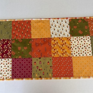 May include: A rectangular patchwork table runner featuring a variety of autumn-themed fabric squares in shades of orange, yellow, burgundy, and olive green. The patterns include pumpkins, corn, leaves, small houses, and geometric grids.