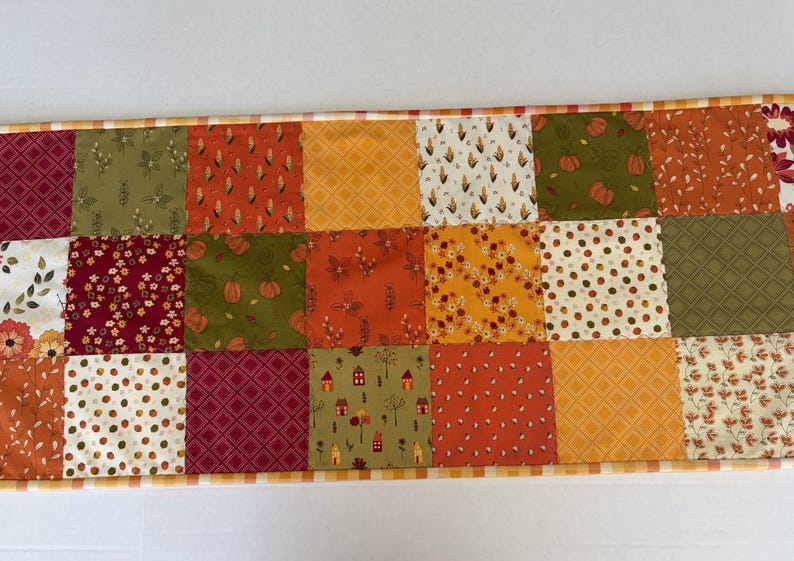 May include: A rectangular patchwork table runner featuring a variety of autumn-themed fabric squares in shades of orange, yellow, burgundy, and olive green. The patterns include pumpkins, corn, leaves, small houses, and geometric grids.