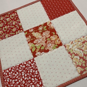 May include: A square patchwork quilt with a red and white color scheme. The quilt is made of various fabric squares, including floral and dotted patterns. The edges are finished with a red and white gingham binding.