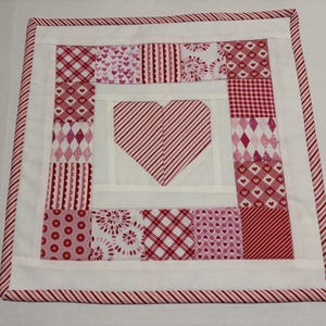 May include: A square quilted placemat with a heart design. The heart is made of red and white striped fabric, surrounded by squares of patterned fabric in shades of red and pink. The border is white with a red and white striped trim.