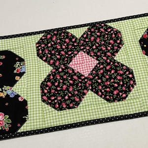 May include: A floral table runner featuring a green gingham background with black and white polka dot trim. The runner has black floral patterned sections with pink and red roses and a pink and white checkered center.