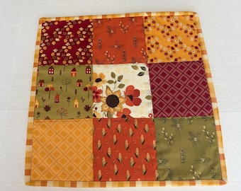 May include: A square patchwork quilt featuring nine distinct fabric panels with autumn-themed patterns including sunflowers, corn stalks, small houses, and geometric shapes, all bordered by a yellow and white checkered fabric trim.