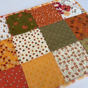 May include: A rectangular fabric table runner featuring a patchwork design of twelve squares with various autumn-themed patterns including pumpkins, corn, flowers, leaves, and small houses in shades of orange, green, yellow, and white with a plaid border.