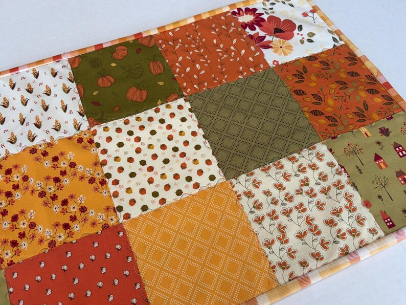 May include: A rectangular fabric table runner featuring a patchwork design of twelve squares with various autumn-themed patterns including pumpkins, corn, flowers, leaves, and small houses in shades of orange, green, yellow, and white with a plaid border.