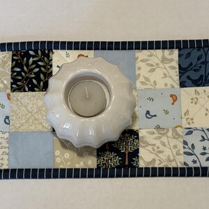 May include: A rectangular quilted table runner featuring a patchwork design of blue, navy, cream, and white fabric squares with patterns of birds, leaves, and script text. A white, scalloped-edge ceramic candle holder sits in the center.