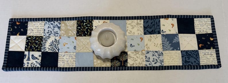 May include: A rectangular quilted table runner featuring a patchwork design of blue, navy, cream, and white fabric squares with patterns of birds, leaves, and script text. A white, scalloped-edge ceramic candle holder sits in the center.