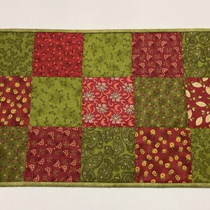 Puede incluir: Camino de mesa acolchado con dise&ntilde;o de patchwork. Los cuadrados alternan entre tonos de rojo y verde, con estampados florales y de cachemira. El camino de mesa est&aacute; rematado con un borde verde.