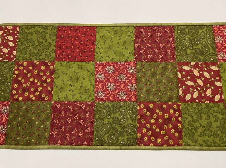Puede incluir: Camino de mesa acolchado con dise&ntilde;o de patchwork. Los cuadrados alternan entre tonos de rojo y verde, con estampados florales y de cachemira. El camino de mesa est&aacute; rematado con un borde verde.