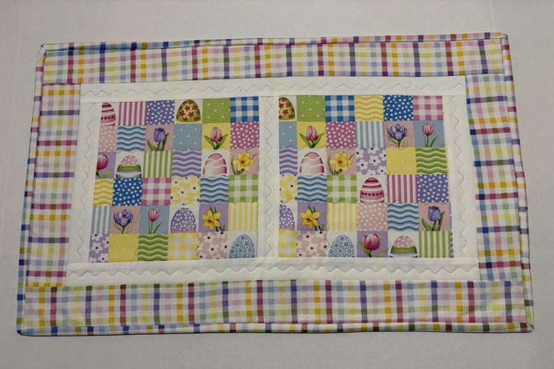 Pode incluir: Esteira de P&aacute;scoa com um design de patchwork. O centro apresenta quadrados com ovos de P&aacute;scoa, flores e padr&otilde;es em cores pastel. Uma borda branca com vieiras emoldura o centro, e uma borda xadrez em tons pastel envolve toda a esteira.