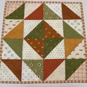May include: A square fabric quilt featuring a patchwork design of triangles in shades of olive green, burnt orange, and cream. The pattern includes various small floral, leaf, and acorn prints, finished with a striped border in brown and white.
