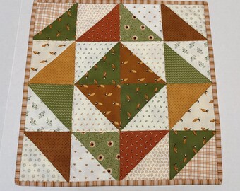 May include: A square fabric quilt featuring a patchwork design of triangles in shades of olive green, burnt orange, and cream. The pattern includes various small floral, leaf, and acorn prints, finished with a striped border in brown and white.