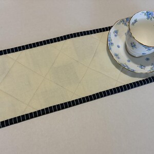 May include: A rectangular quilted table runner in cream fabric with a diamond-stitched pattern and a dark blue and white pinstriped border. A white porcelain teacup and matching saucer with a delicate blue floral pattern rest on the runner.