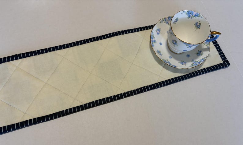 May include: A rectangular quilted table runner in cream fabric with a diamond-stitched pattern and a dark blue and white pinstriped border. A white porcelain teacup and matching saucer with a delicate blue floral pattern rest on the runner.