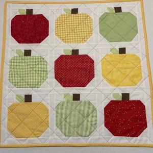 May include: A quilted wall hanging featuring nine apple shapes in red, yellow, and green fabrics. Each apple has a brown stem and green leaf. The apples are arranged in a 3x3 grid on a white background with a yellow border.
