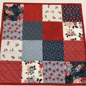 May include: A patchwork quilt with a red border. The quilt is composed of sixteen squares in various patterns, including floral, polka dots, and geometric designs. The color palette includes red, navy, light blue, and white.