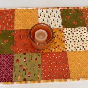 May include: A rectangular patchwork table runner featuring various fabric squares in shades of orange, green, red, and cream, decorated with patterns of pumpkins, corn, houses, and leaves, with a glass candle holder placed in the centre.