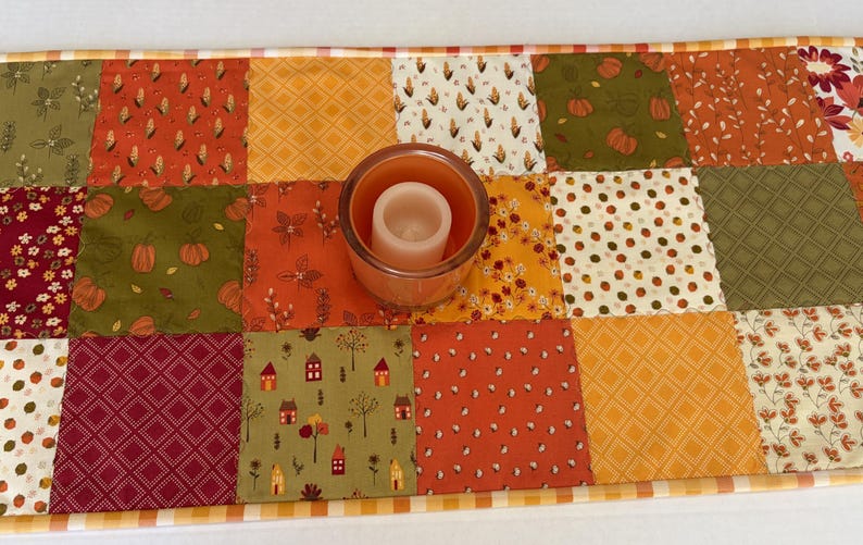 May include: A rectangular patchwork table runner featuring various fabric squares in shades of orange, green, red, and cream, decorated with patterns of pumpkins, corn, houses, and leaves, with a glass candle holder placed in the centre.