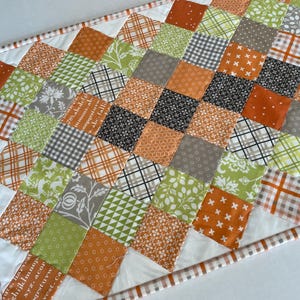 May include: A quilted table runner with a patchwork design. The runner features squares in shades of orange, green, gray, and black, with various patterns like plaid, floral, and geometric shapes. The edges are finished with a white border and a patterned trim.