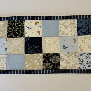 May include: A rectangular quilted table runner featuring a patchwork design of squares in shades of navy blue, light blue, cream, and white. The fabric squares display patterns of birds, branches, and cursive script text, finished with a dark blue striped border.