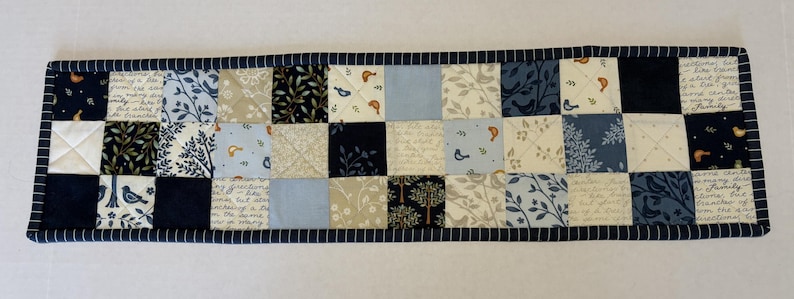 May include: A rectangular quilted table runner featuring a patchwork design of squares in shades of navy blue, light blue, cream, and white. The fabric squares display patterns of birds, branches, and cursive script text, finished with a dark blue striped border.