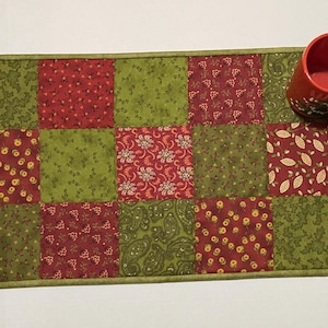 Puede incluir: Un camino de mesa acolchado rectangular con un dise&ntilde;o de patchwork en rojo y verde. El camino de mesa presenta cuadrados con patrones florales y de hojas. Dos tazas rojas con dise&ntilde;os florales blancos est&aacute;n encima.