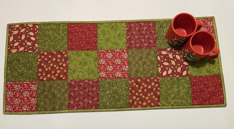 Puede incluir: Un camino de mesa acolchado rectangular con un dise&ntilde;o de patchwork en rojo y verde. El camino de mesa presenta cuadrados con patrones florales y de hojas. Dos tazas rojas con dise&ntilde;os florales blancos est&aacute;n encima.