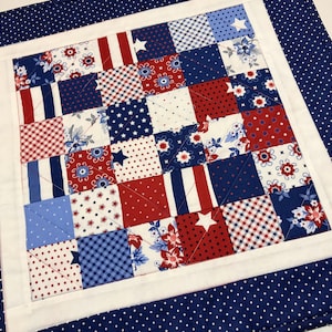 May include: A square patchwork quilt with a patriotic theme. The center features a grid of red, white, and blue squares with various patterns, including stars, stripes, and floral designs. A white border surrounds the patchwork, and a navy blue polka dot fabric frames the quilt.