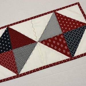 May include: A rectangular quilted table runner with a white center and a red and white patterned border. The center features alternating red, white, and blue triangles with various patterns, including stars, gingham, and floral designs.